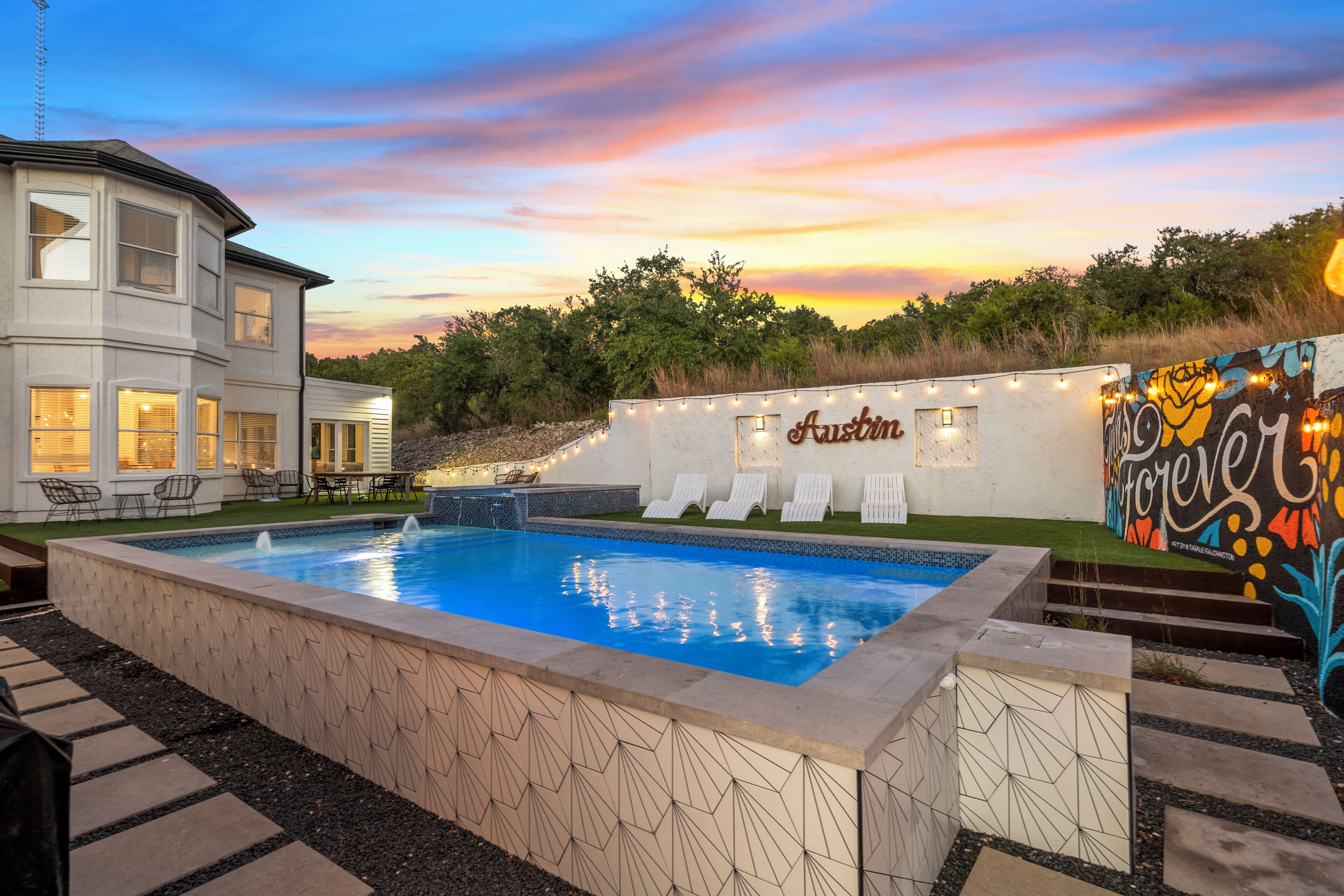 A swimming pool with a geometric tile design, surrounded by a landscaped area with a white building and a mural.