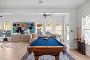 Una mesa de billar en una habitación con un ventanal grande, un ventilador de techo y una pared decorada con varios sombreros.