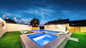 A rectangular swimming pool with a tiled edge, surrounded by artificial grass, under a night sky with string lights. - Luxury 7BR - Heated Pool, Hot Tub, Pet Friendly (Austin)