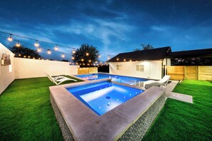 A rectangular swimming pool with a tiled edge, surrounded by artificial grass, under a night sky with string lights.
