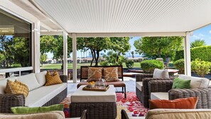 A covered outdoor seating area with wicker furniture, a central coffee table, and a view of a landscaped garden and distant buildings.