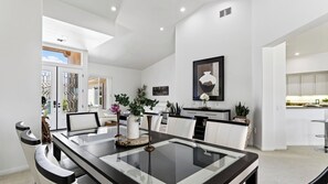 A modern dining area with a glass-top table, white upholstered chairs, and a kitchen in the background.