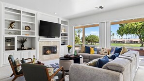 A modern living room with a fireplace, television, and large windows overlooking a landscaped area. - Palm & Par by Avantstay Golf Views + Patio BBQ Mins to Indian Wells (Palm Desert)