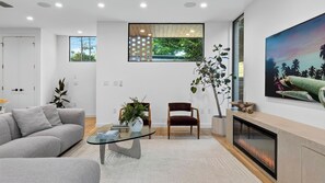 Una sala moderna con sofá, una mesa de centro de vidrio, chimenea y un ventanal con vistas al follaje.