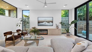 Una sala de estar moderna con una mesa de centro de vidrio, asientos cómodos, una chimenea y ventanas amplias con vistas a follaje.