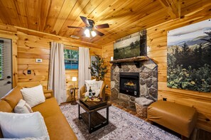 A cozy log cabin interior with a stone fireplace, a sofa, a coffee table, and a painting of a landscape. - Havenheart Smoky Mtns Romantic Treehouse (Sevierville)