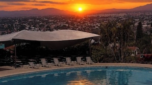Una piscina con sillones de descanso y sombrillas, con vista a un atardecer sobre el paisaje urbano y las montañas.