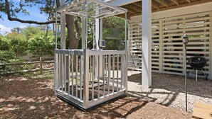 A metal structure with a white frame, possibly a gazebo or enclosure, in a garden area with trees and a barbecue grill in the background. - Coastal Escape, Pool, Hot Tub & Resort Amenities (Currituck County)