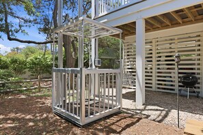 Eine Metallkonstruktion mit weißem Rahmen, möglicherweise ein Gartenpavillon oder eine Umzäunung, in einem Gartenbereich mit Bäumen und einem Grill im Hintergrund.