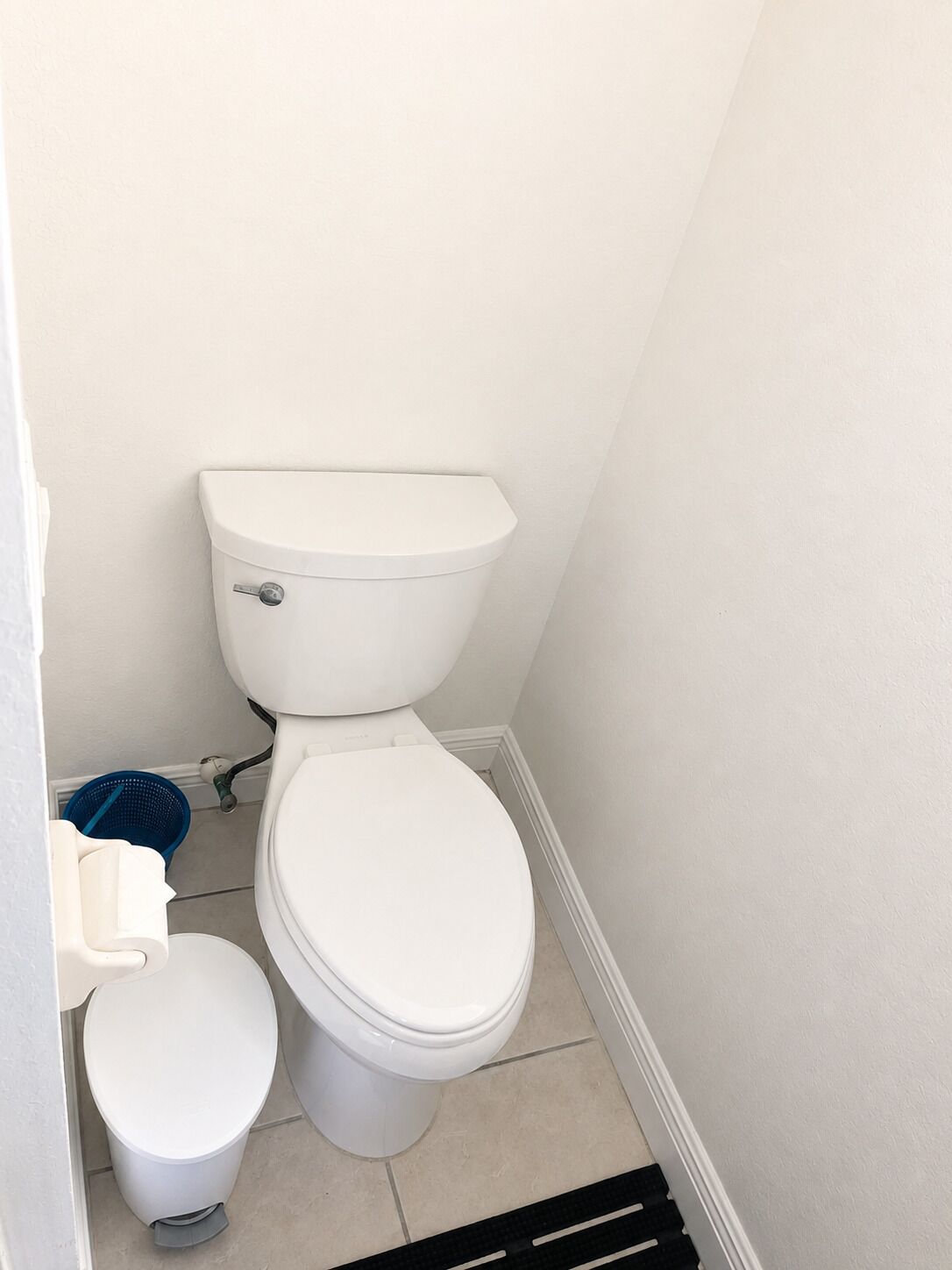 Un inodoro estándar con tanque y asiento, un pequeño cubo de basura y una cesta azul para limpieza.