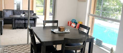 A dining area with a table and chairs, a ceiling light fixture, and a view of a swimming pool through a window.