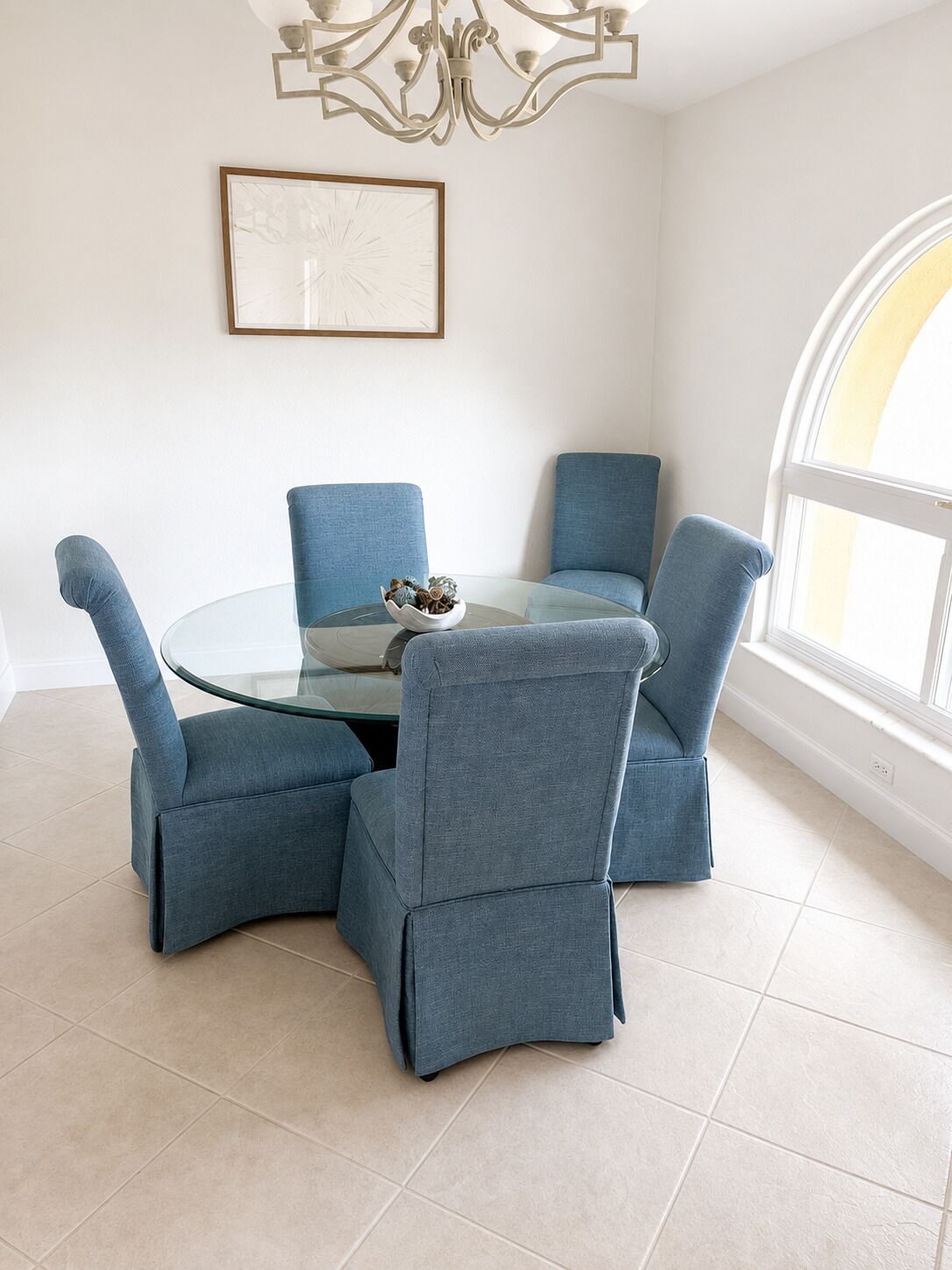 A dining area with a glass table surrounded by four blue upholstered chairs, a chandelier, and a framed artwork on the wall.