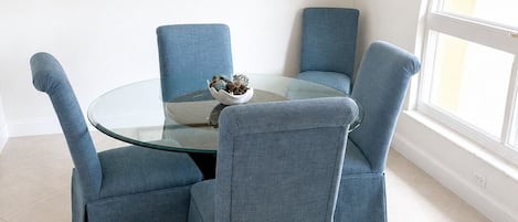 A dining area with a glass table surrounded by four blue upholstered chairs, a chandelier, and a framed artwork on the wall.