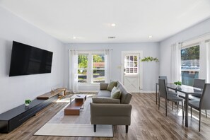 Una sala de estar moderna con un televisor de pantalla plana, un sofá, una mesa de comedor con sillas y ventanas amplias que ofrecen vistas al exterior.
