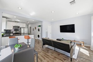 A modern apartment with a kitchen, dining area, and living room. It includes a dining table with chairs, a sofa, a TV mounted on the wall, and a washing machine in the background.