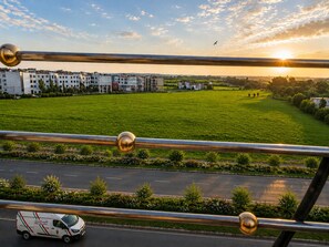 A view of a green field with a building complex in the background, a road with a vehicle, and a clear sky with the sun setting.