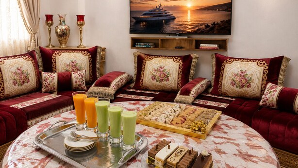 A living room with a round table, sofas, a tray with drinks and pastries, and a painting of a sunset over water.