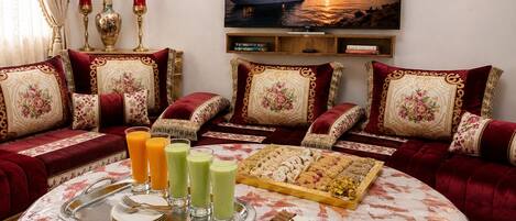 A living room with a round table, sofas, a tray with drinks and pastries, and a painting of a sunset over water.