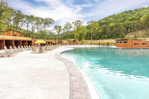 A swimming pool with lounge chairs and umbrellas, surrounded by wooden cabins and a forested hillside.