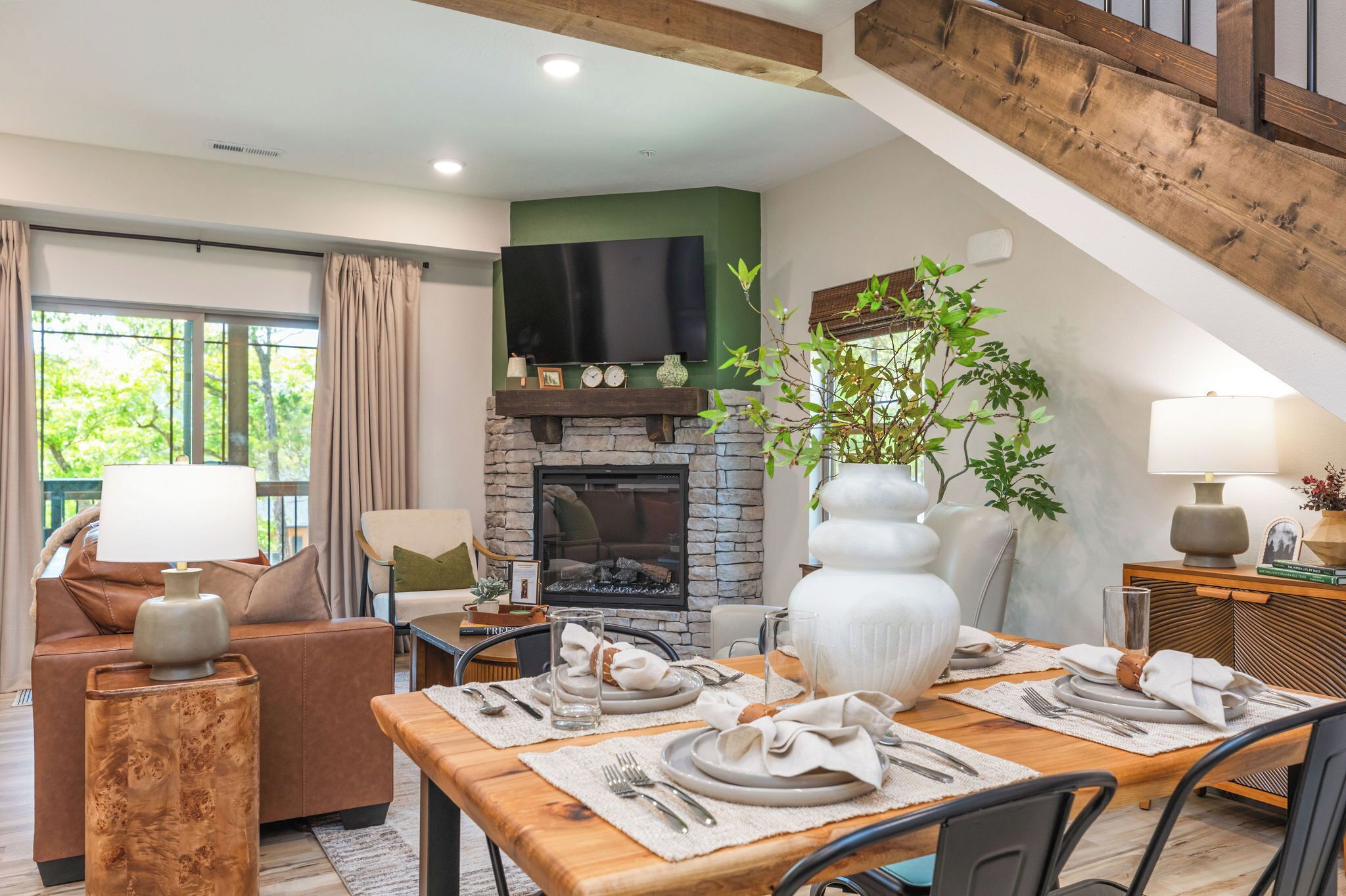 A dining area with a wooden table set for two, a stone fireplace, and a green wall with a flat-screen TV above it.