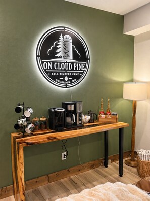 A room with a wall-mounted sign reading 'ON CLOUD PINE,' a coffee station with a coffee maker and mugs, and a lamp on a side table.