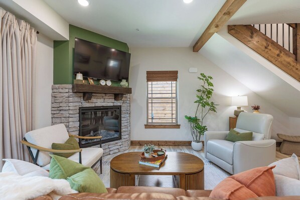 A cozy living room with a stone fireplace, a flat-screen TV mounted above it, and a comfortable seating area with a sofa and chairs.
