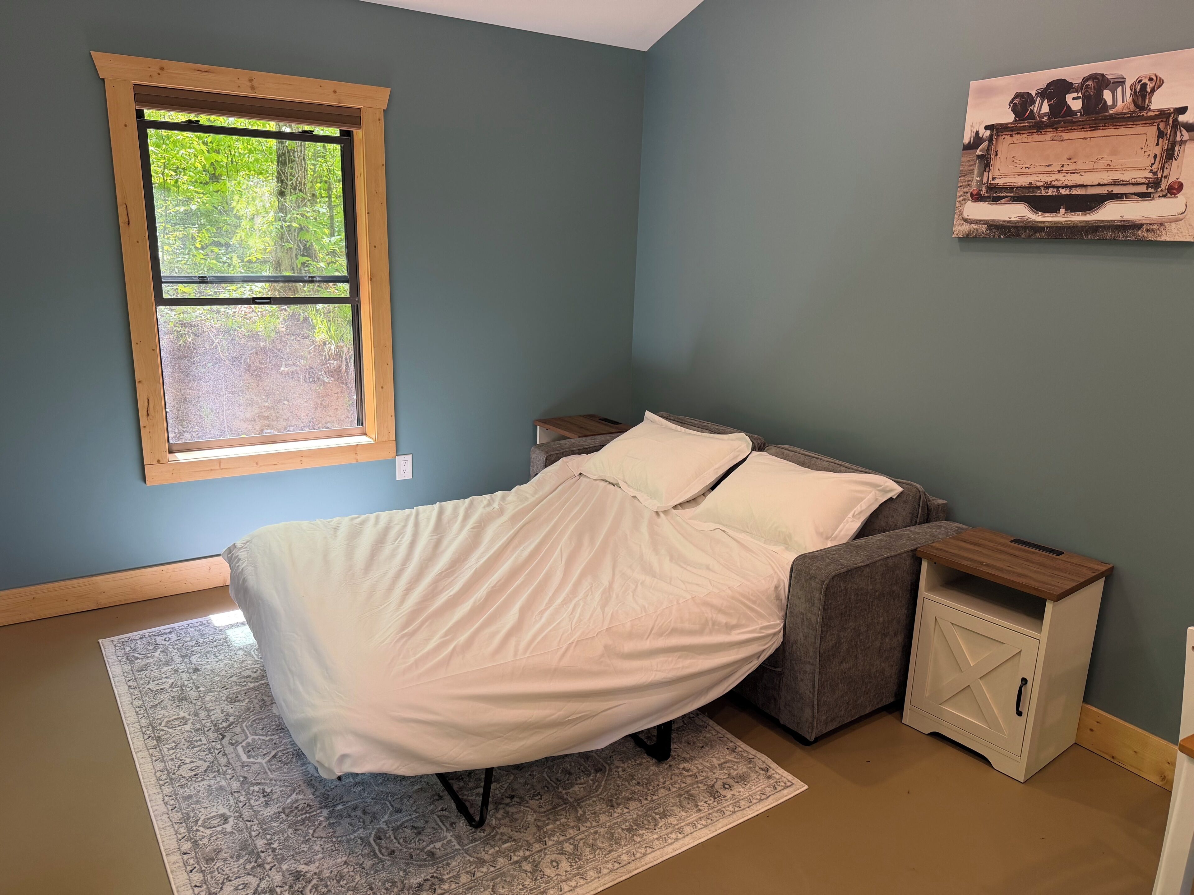 A bedroom with a bed, a window, a nightstand, a cabinet, and a wall-mounted picture.