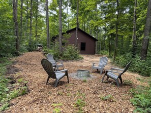 A cabin surrounded by a forest with outdoor seating and a fire pit.