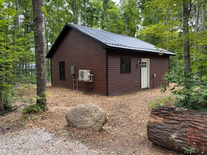 Eine kleine, eingeschossige Holzhütte mit einem Metalldach, umgeben von Bäumen und einem Schotterweg.