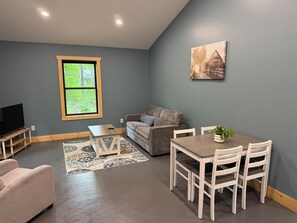 A living room with a gray sofa, a coffee table, a TV on a stand, a dining table with chairs, and a window with a view of trees.