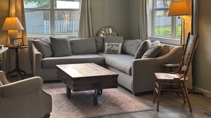 Een woonkamer met een hoekbank, een houten salontafel, een houten stoel, een lamp en een decoratief wandobject.