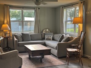 Ein Wohnzimmer mit einem Ecksofa, einem Holztisch, einem Holzstuhl, einer Lampe und einer Wanddekoration.