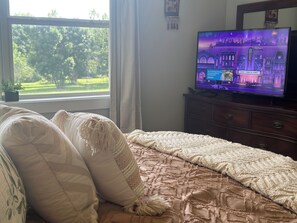 Ein Schlafzimmer mit einem Bett, einem Fernseher auf einer Kommode und einem Fenster mit Blick auf Bäume.
