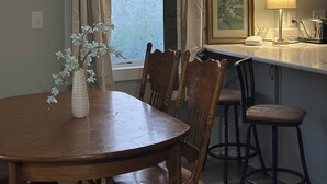 Een eethoek met een houten tafel en stoelen, een vaas met bloemen, een raam met gordijnen en een keuken met een lamp.