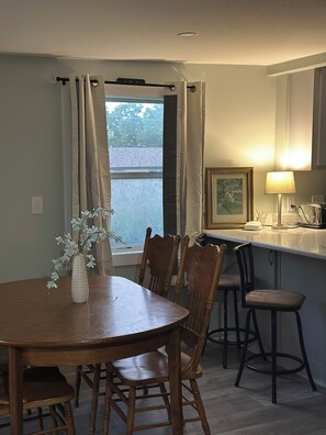 Ein Essbereich mit Holztisch und Stühlen, einer Vase mit Blumen, einem Fenster mit Vorhängen und einer Küchenzeile mit einer Lampe.