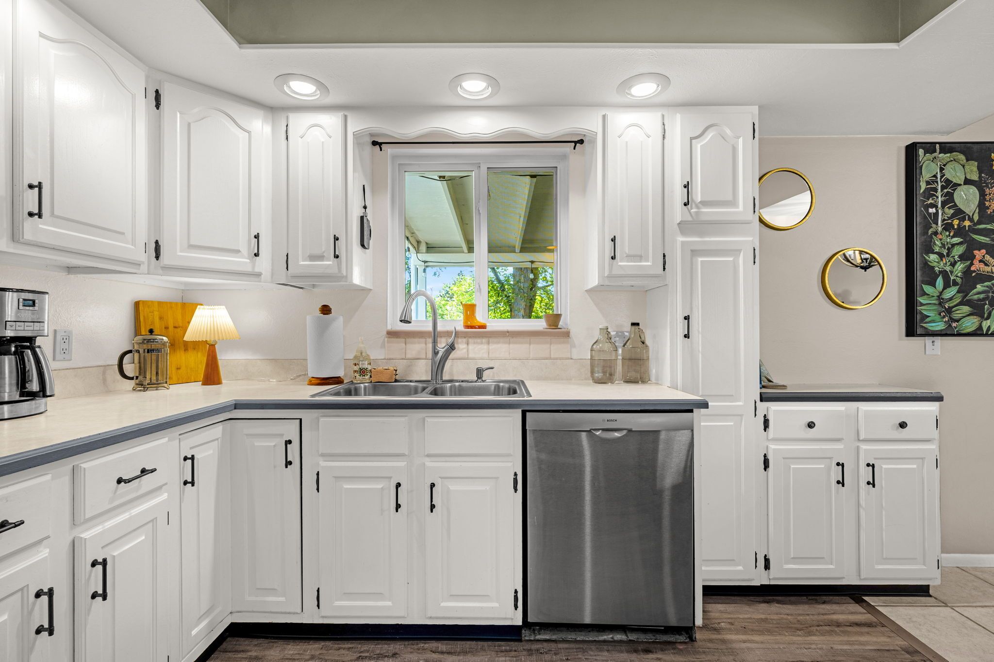 A kitchen with white cabinets, a stainless steel dishwasher, and a window with a view of greenery.