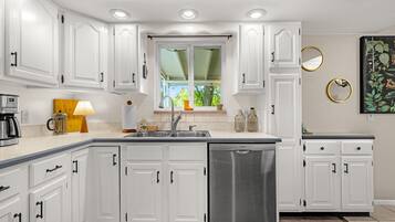 A kitchen with white cabinets, a stainless steel dishwasher, and a window with a view of greenery.