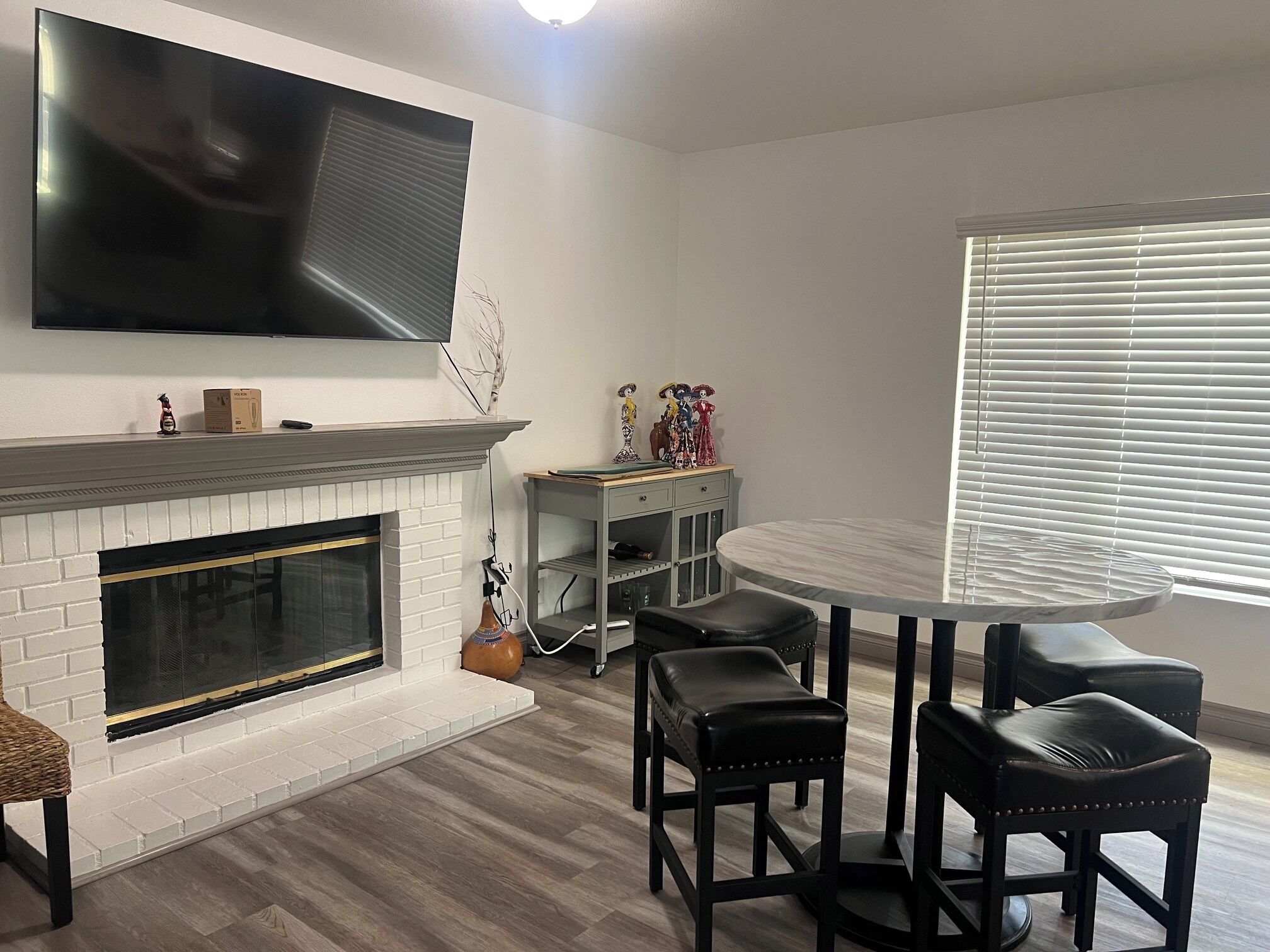 A modern living room with a fireplace, a flat-screen TV mounted on the wall, a round dining table with chairs, and a window with blinds.