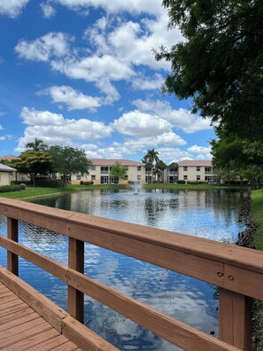A wooden deck overlooking a calm pond with a fountain, surrounded by buildings with a uniform architectural style. - Spacious 3-bedroom condo with pool/fitness center in lovely Fort Myers (Fort Myers)