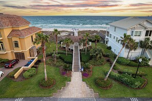 A coastal property with a staircase leading to a deck overlooking the ocean, surrounded by palm trees and well-maintained gardens. - SeaHaven @ Laguna Key- 6BR "Almost Beachfront" Home, Pool, Tiki Bar, Game Garage (Gulf Shores)