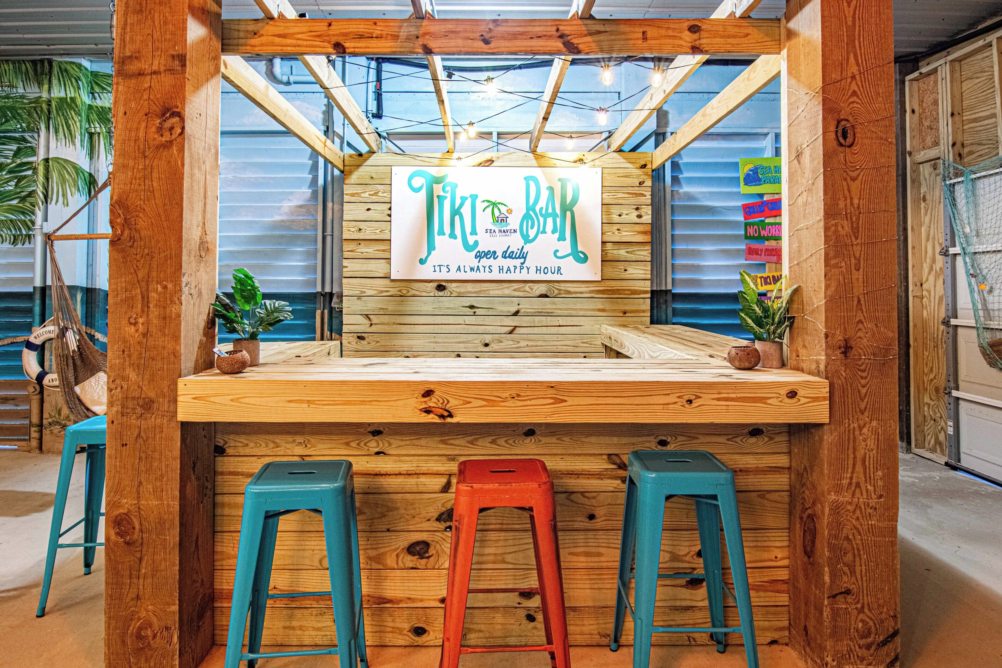 Un comptoir en bois, sur lequel on peut lire les inscriptions « Tiki Bar » et « C’est toujours l’heure de l’apéro ». Il y a trois tabourets devant le comptoir.