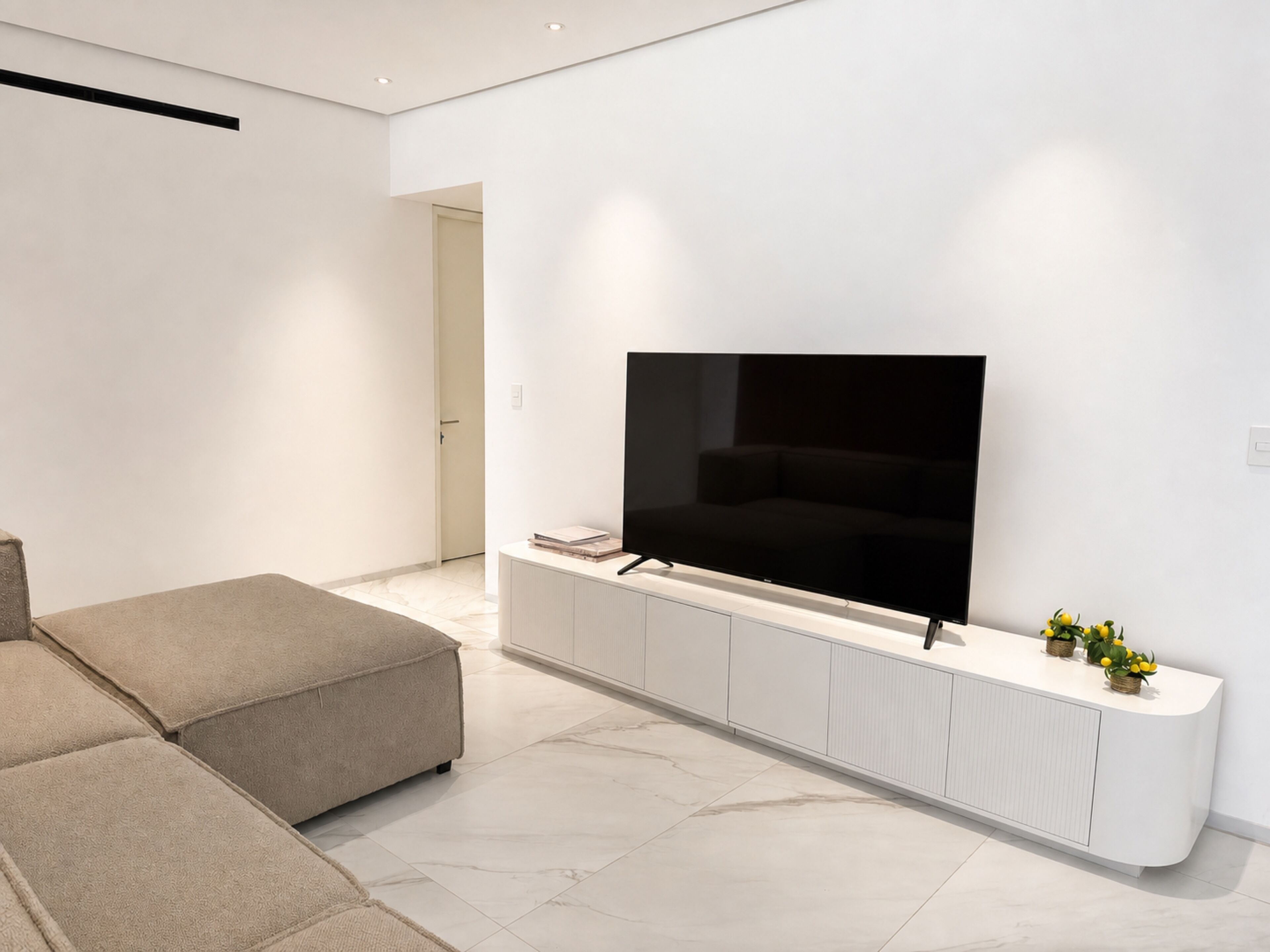 Un salon moderne avec un téléviseur à écran plat fixé sur une console blanche, un canapé beige et un design minimaliste.