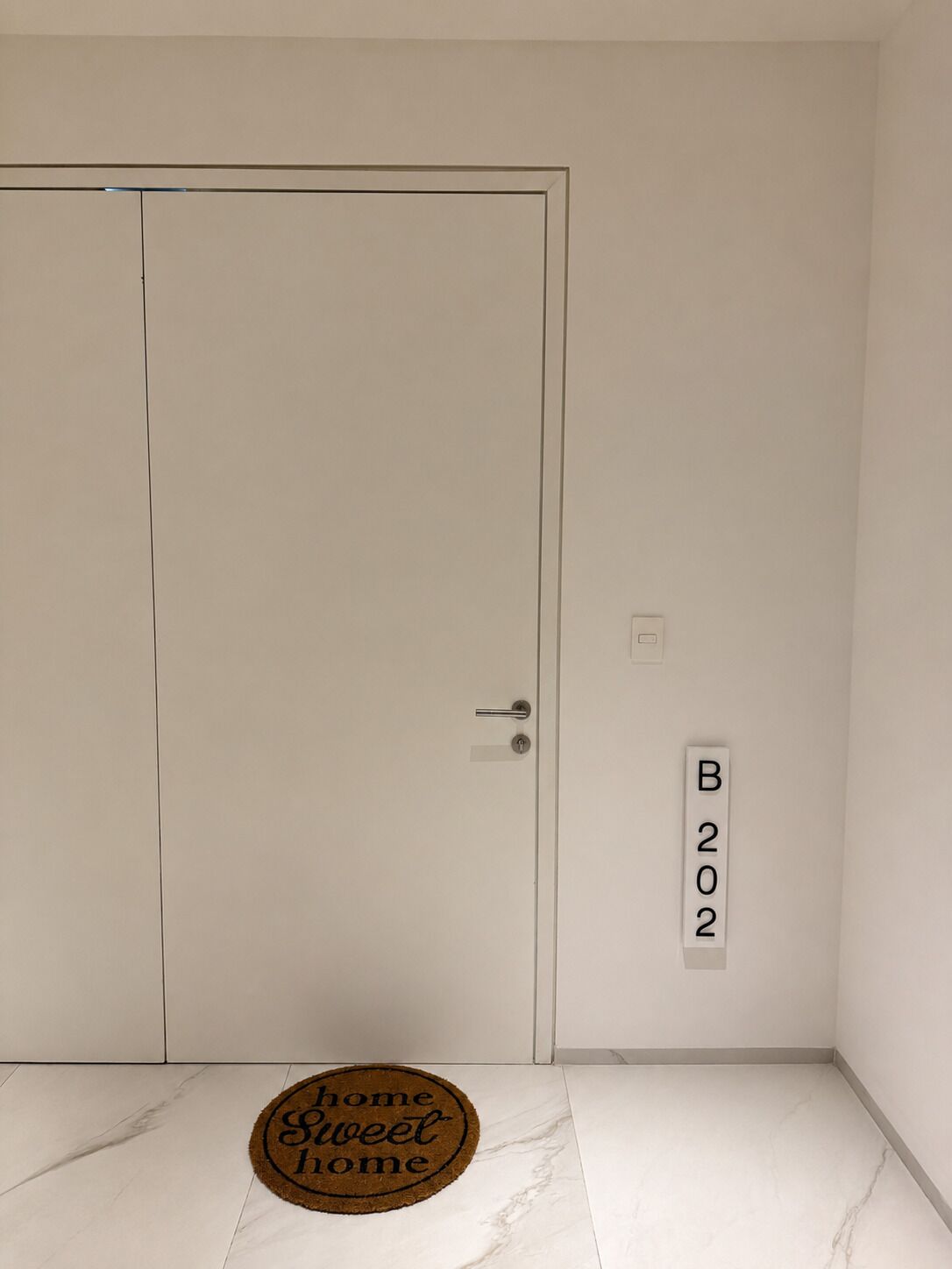 A room with a white door, a doormat with 'home sweet home' text, and a wall-mounted sign with 'B202'.