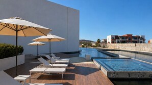Un área de piscina en la azotea con sillones de descanso y sombrillas, un cielo azul despejado y vistas a edificios en la distancia.