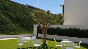Un área de estar moderna al aire libre con sillas y mesas blancas, un árbol y un edificio de fachada blanca y un seto verde.
