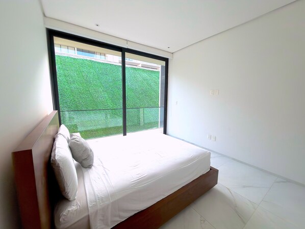 A bedroom with a large window, a bed, and a view of a green outdoor area.