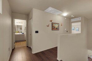 A hallway with a wooden floor, a bathroom with a vanity and mirror, and a wall with a framed picture and wall-mounted lights.