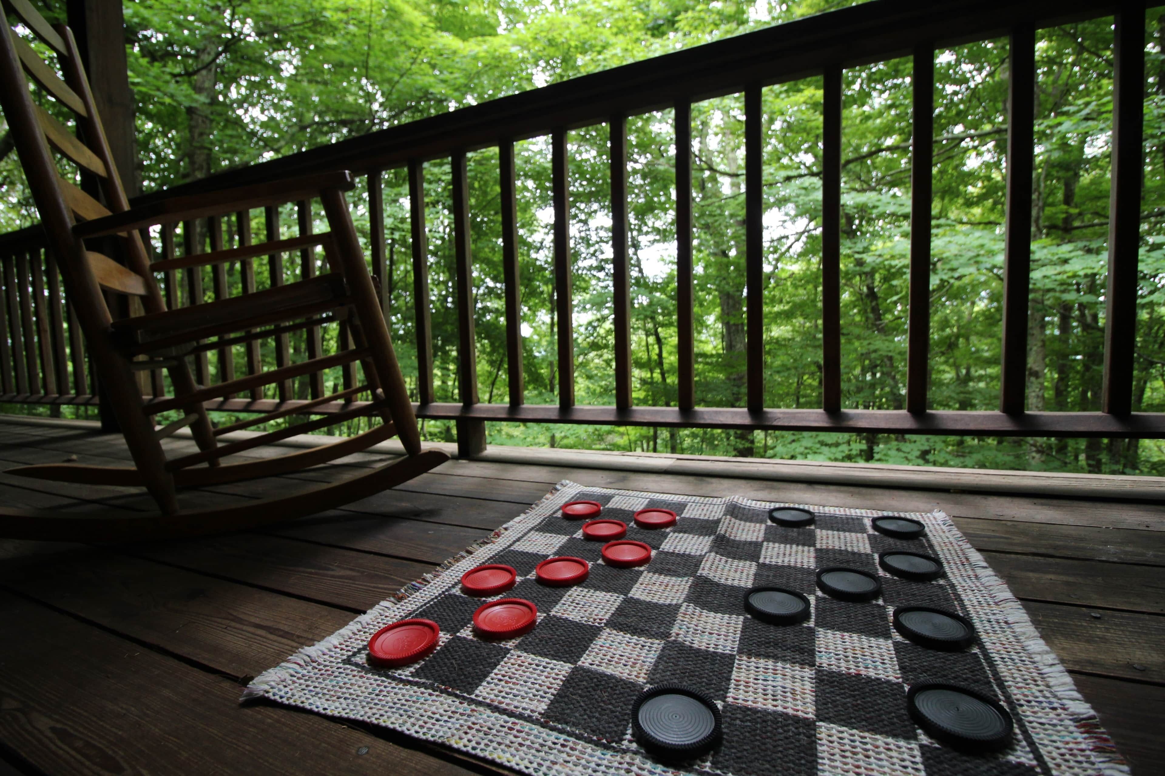 Un jeu de société à damier est installé sur une terrasse en bois dotée d’une rambarde, offrant une vue sur une forêt.