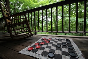Un jeu de société à damier est installé sur une terrasse en bois dotée d’une rambarde, offrant une vue sur une forêt.