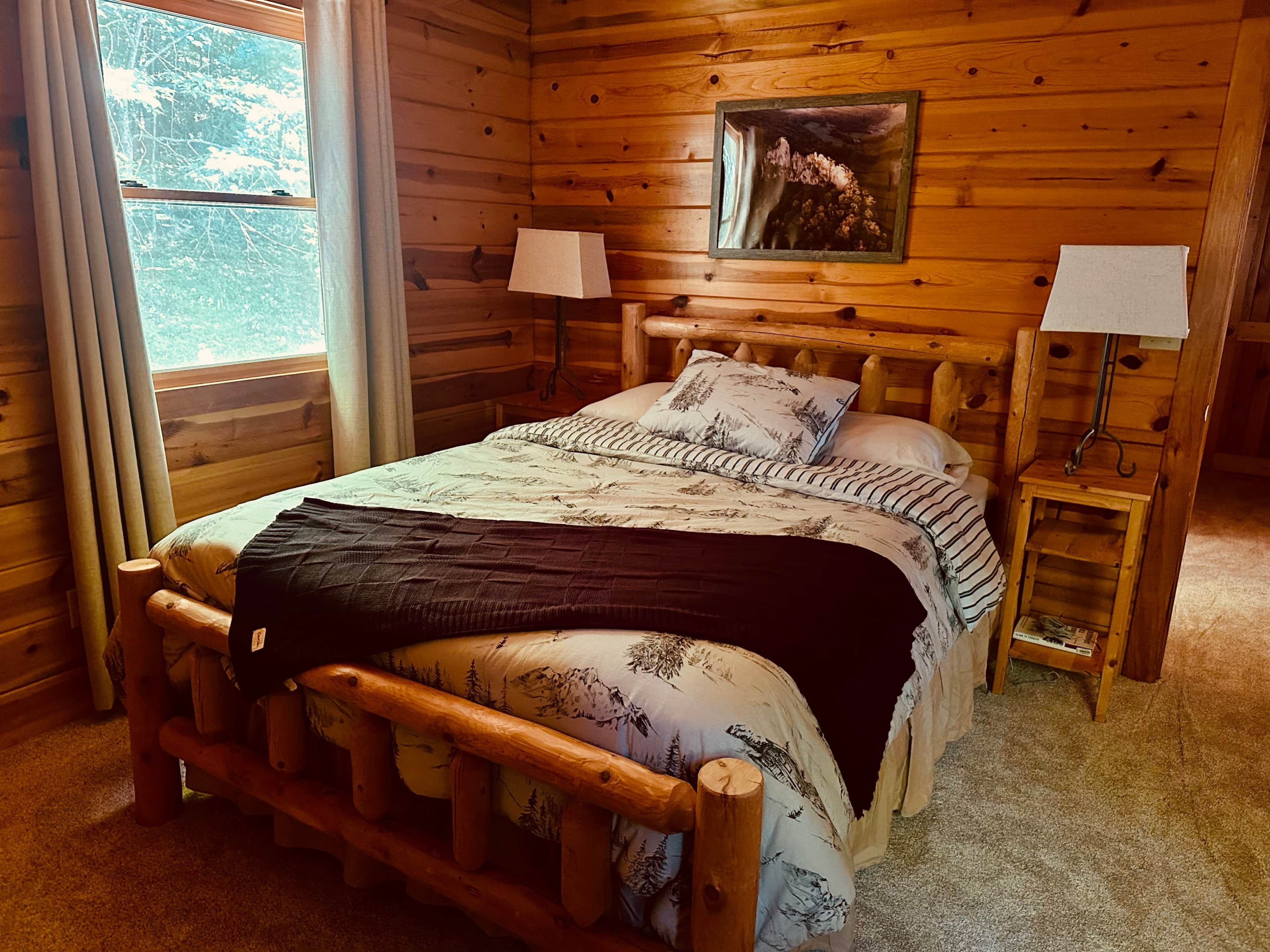 Une chambre à coucher dans une cabane en bois, avec un lit, deux lampes de chevet, une fenêtre et un tableau accroché au mur.
